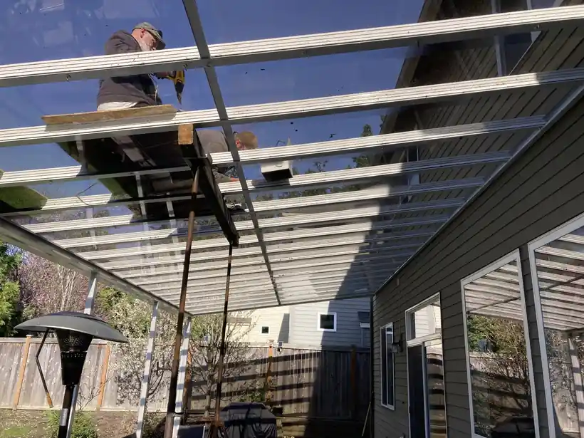 Two installers working on the glazing bars of a clear glass patio cover, seen from underneath